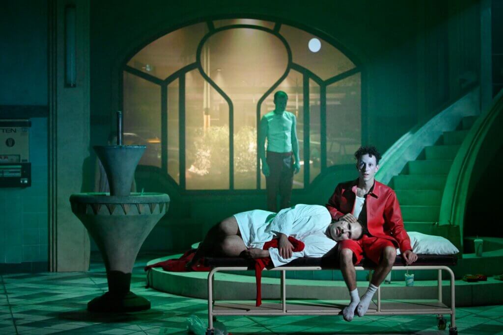 „Double Serpent” von Sam Max am Staatstheater Wiesbaden. In dämmerigen Licht sitzt ein Mann auf einem Bettgestell, neben ihm liegt ein Mann mit seinem Kopf auf seinem Schoß. Im Hintergrund steht ein Mann und schaut nach unten. Dahinter ist eine große Fensterfront.