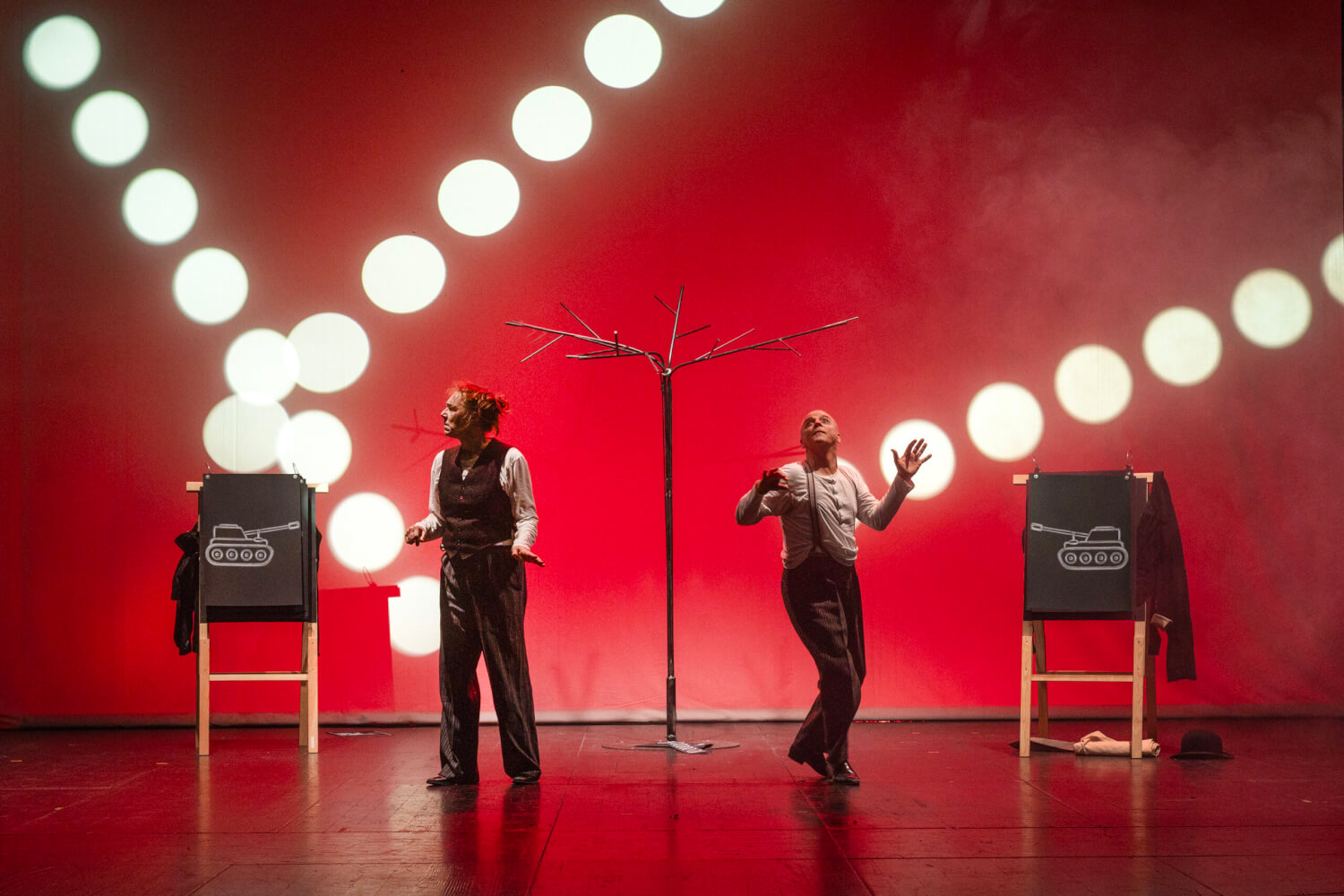 Aufführungsfoto von „Game of Clowns“ von Erkan Uyanıksoy der Bremer Shakespeare Company. Vor einem rot erleuteten Hintergrund mit hell leuchtenden Kreisen steht eine Frau und ein Mann. Auf zwei Tafeln sind jeweils ein mit Kreide gemalter Panzer zu sehen. Der Mann schaut gespannt nach oben, die Frau schaut zur Seite.