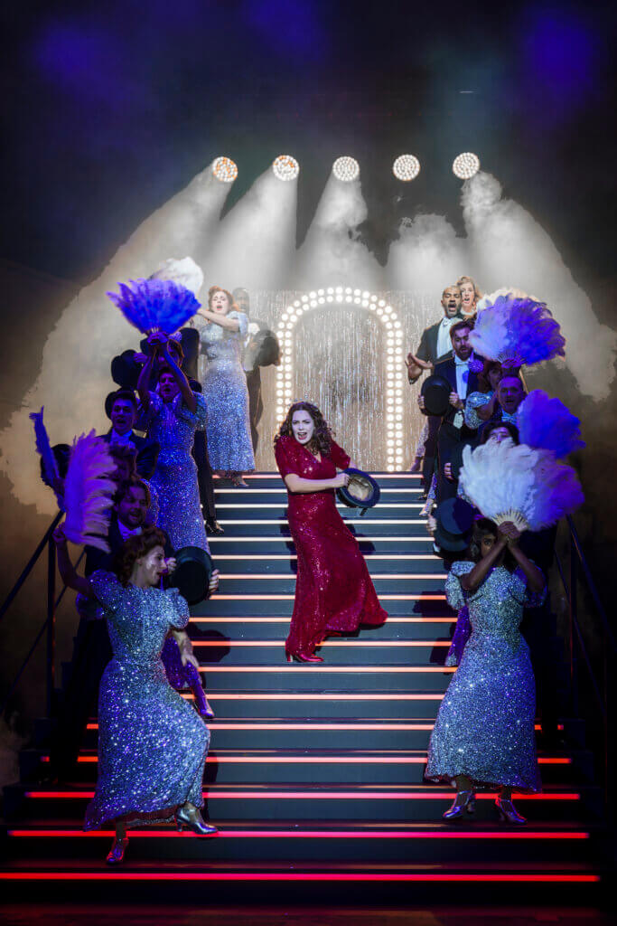Aufführungsfoto von „Grand Finale. Ein Geistermusical“ von Christoph Israel am Theater Basel. Eine Frau im rot glitzernden Abendkleid geht eine Showtreppe runter. Sie wird flankiert auf der Treppe von Frauen und Männern mit Federboas und Showroben.