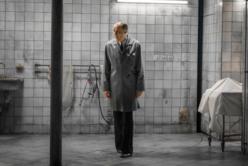 Aufführungsfoto von „Grand Finale – Ein Geistermusical“ von Christoph Israel am Theater Basel. Ein Mann steht in einem grauen Mantel starr vor einer grauen Fliesenwand.