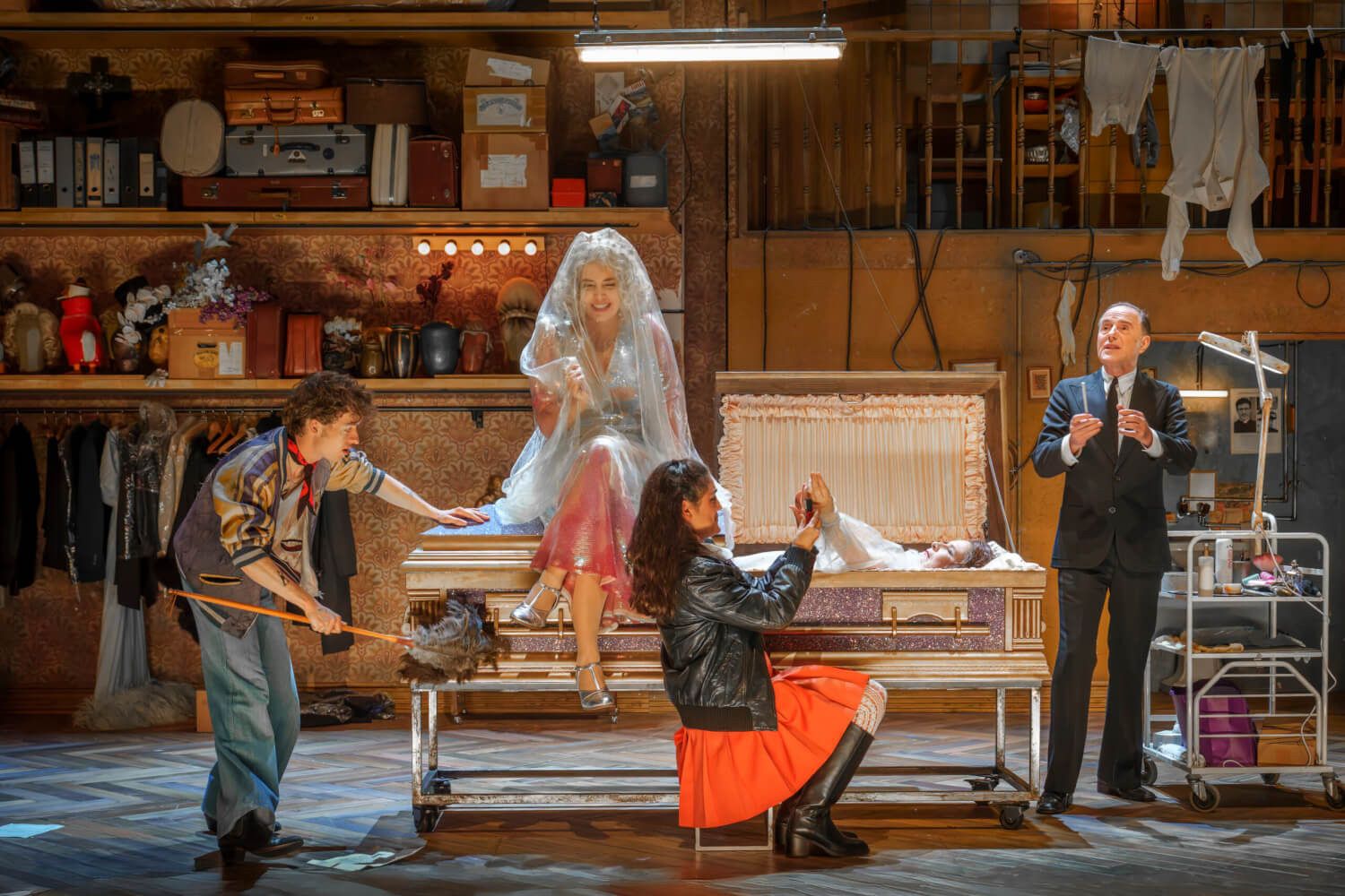 Aufführungsfoto von „Grand Finale – Ein Geistermusical“ von Christoph Israel am Theater Basel. Eine Frau mit schwarzer Lederjacke und rotem Rock sitzt neben einem offenen Sarg und macht der darin ruhenden Frau die Nägel. Daneben sitzt eine andere Frau auf dem Sarg. Ein junger Mann wedelt am Fußende des Sargs mit einem Staubwedel. Hinter dem Kopfende des Sargs steht ein älterer Mann in einem schwarzen Anzug.