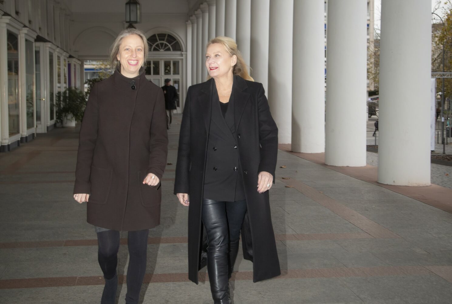 Dorothea Hartmann lächelnd (links) und Beate Heine (rechts) ganz in schwarz bekleidet, laufen frontal auf die Kamera zu, im Außengang des Staatstheater Wiesbaden.