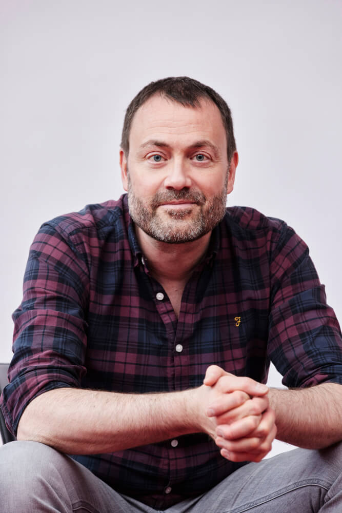 Intendant des schauspiel erlangen, Jonas Knecht, der ein dunkelrot kariertes Hemd an hat und die Ellenbogen auf den Knien abstützt, die Hände sind gefaltet.