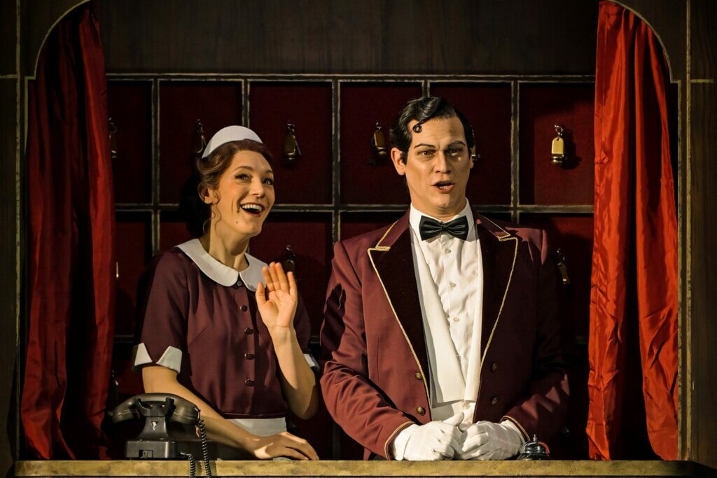 Das Personal steht an der Hotelrezeption. Im Hintergrund sind die Schlüssel zu den Zimmern aufgehängt.