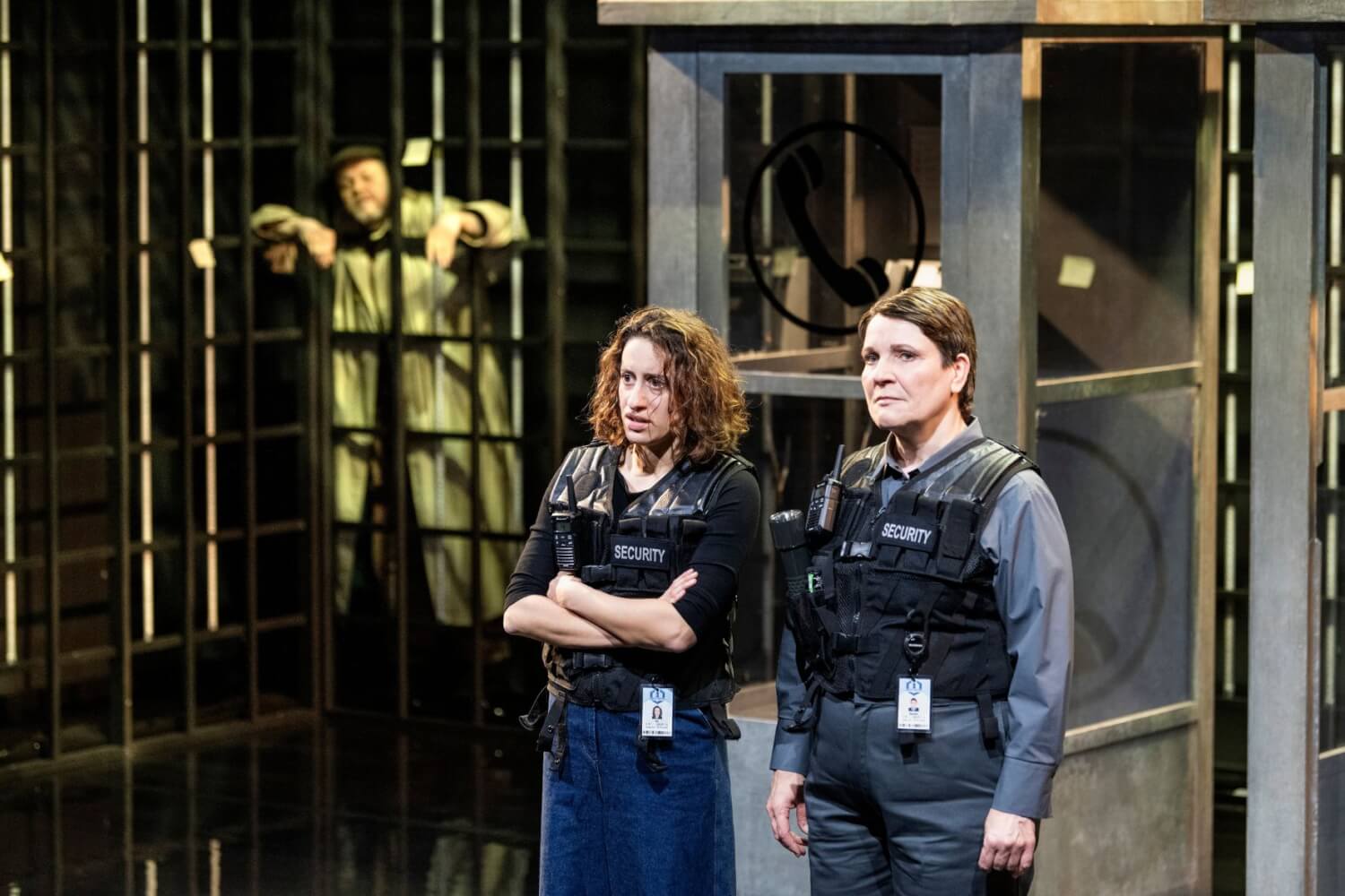 Aufführungsfoto von „Mysteryland“ von Sarah Kilter am Theater Aachen. Zwei Frauen mit schusssicheren Westen und der Aufschrift Security stehen nebeneinander. Im Hintergrund links reckt ein älterer Mann seine Arme durch Gitterstäbe.