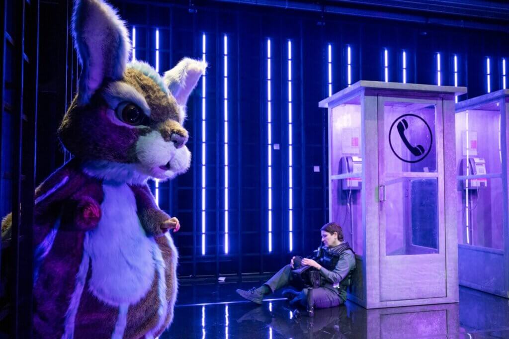 Aufführungsfoto von „Mysteryland“ von Sarah Kilter am Theater Aachen. Links vorne steht ein überdimensionierter Hase. Weiter hinten lehnt eine Frau in Polizeiuniform an einer Telefonzelle.