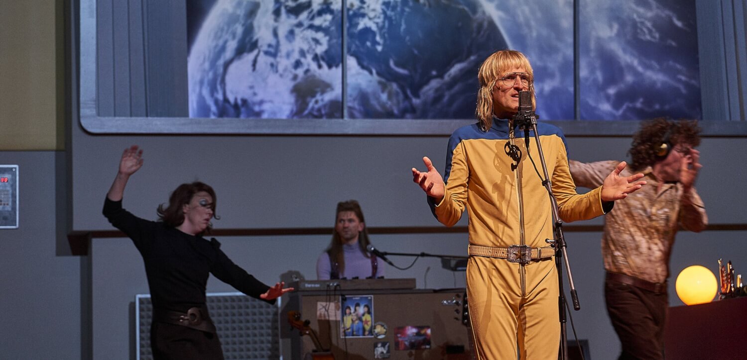 Aufführungsfoto von „Die Nibelungen“ von Bürk und Sienknecht. Ein Mann im ockerfarbenen Anzug steht an einem Mikrofon. Im Hintergrund drei weitere Personen.