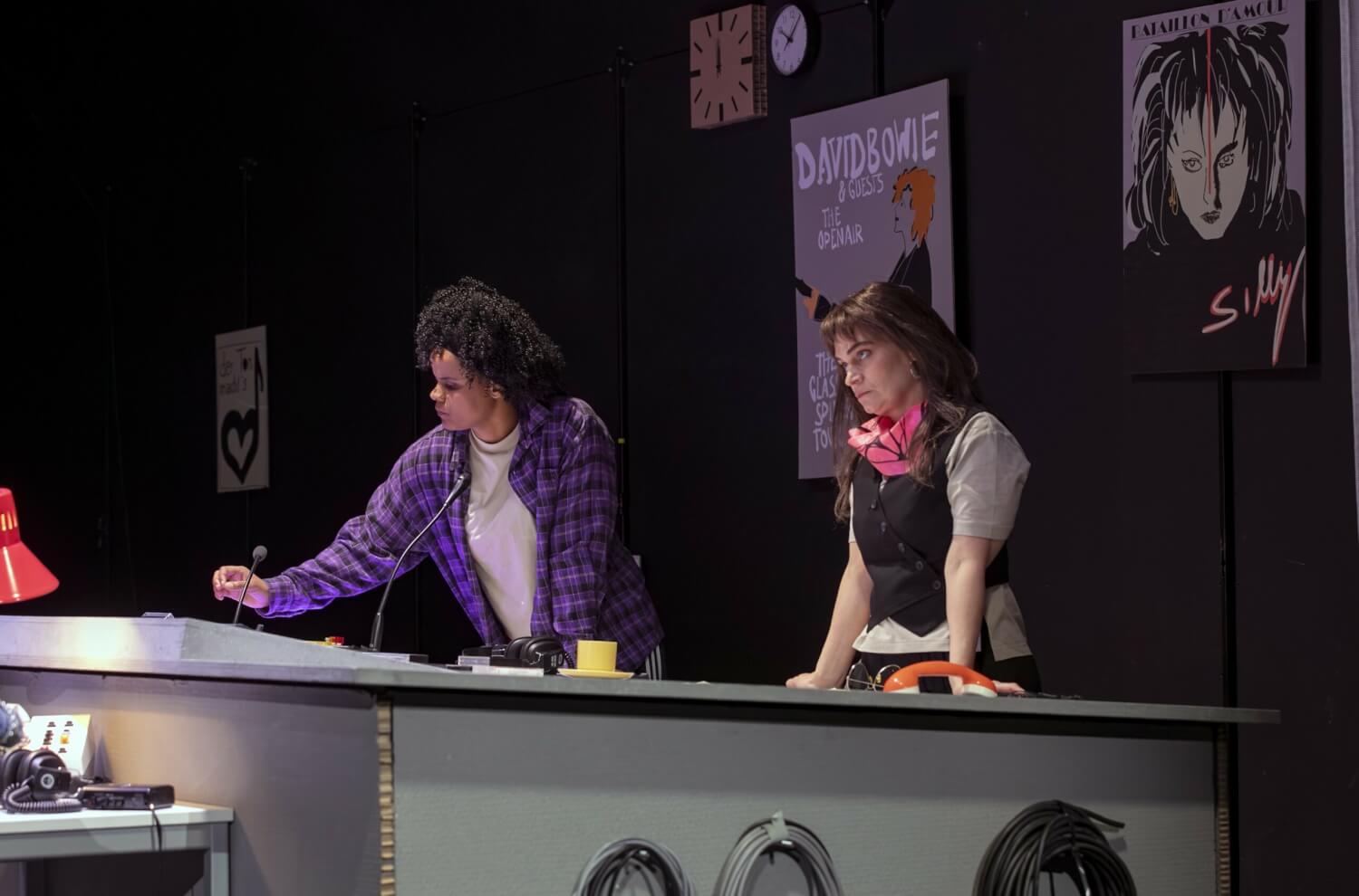 Aufführungsfoto von „On Air On Fire“ von Marion Brasch am Theater an der Parkaue. Zwei Frauen stehen an einem Tonpult und schauen konzentriert. Im Hintergrund an der Wand hängen zwei Uhren und Plakate von David Bowie und Silly.