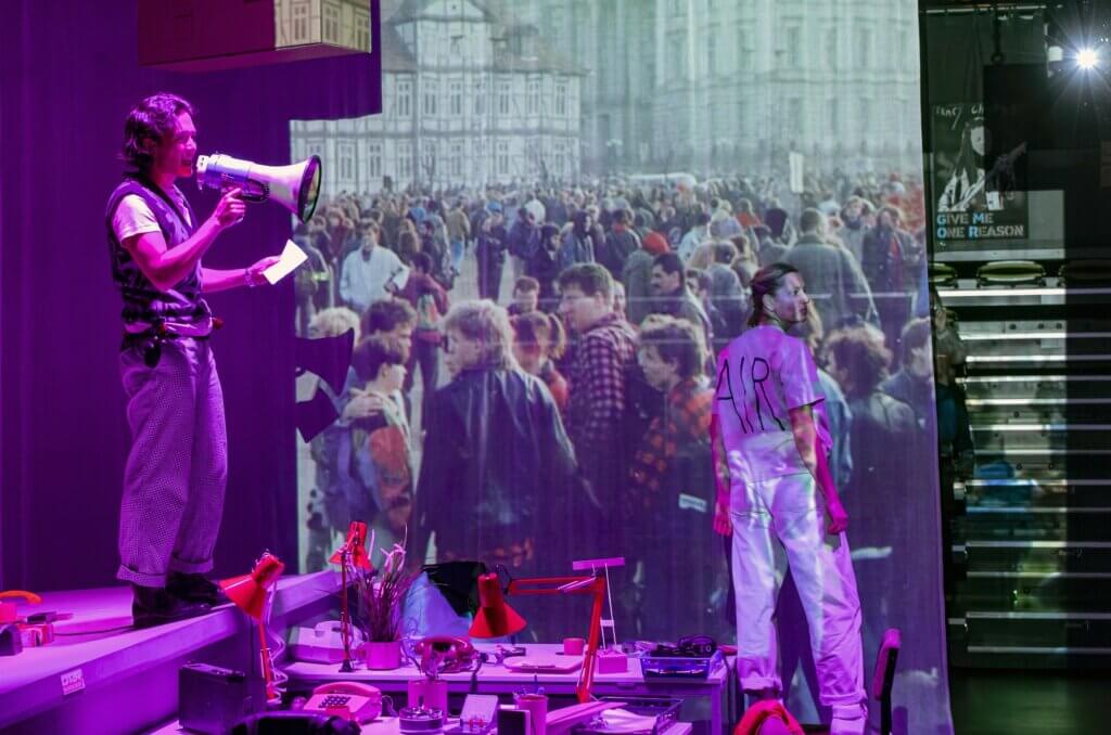 Aufführungsfoto von „On Air On Fire“ von Marion Brasch am Theater an der Parkaue. Ein Mann steht links auf einem Pult und spicht in ein Megaphon, das er in der rechten Hand hält, in der linken hat er einen Zettel. Vor einer Leinwand, auf der eine Menschenmenge im Jahr 1989 zu sehen ist, steht eine Frau und schaut nach rechts.