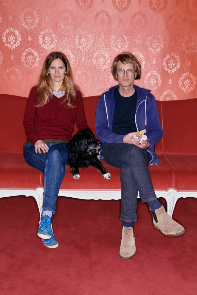Barbara Bürk und Clemens Sienknecht sitzen nebeneinander auf einem roten Plüschsofa, vor einer hellroten Tapete und auf einem roten Samtteppich. Die Beine haben sie synchron übereinandergeschlagen. Zwischen ihnen liegt ein schwarzer Hund. Sienknecht hält ein angebissenes Brötchen in der Hand.