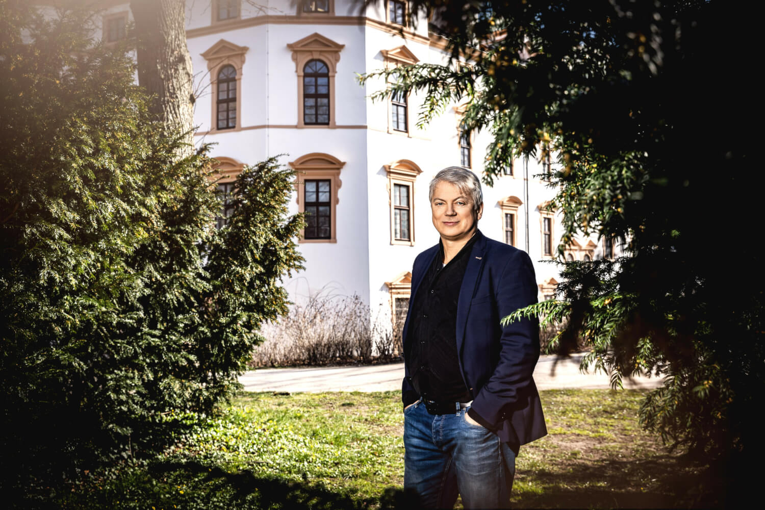 Intendant des Schlosstheater Celle, Andreas Döring. Er steht zwischen zwei Tannen auf einer Wiese. Im Hintergrund die Fassade des Schlosstheaters.