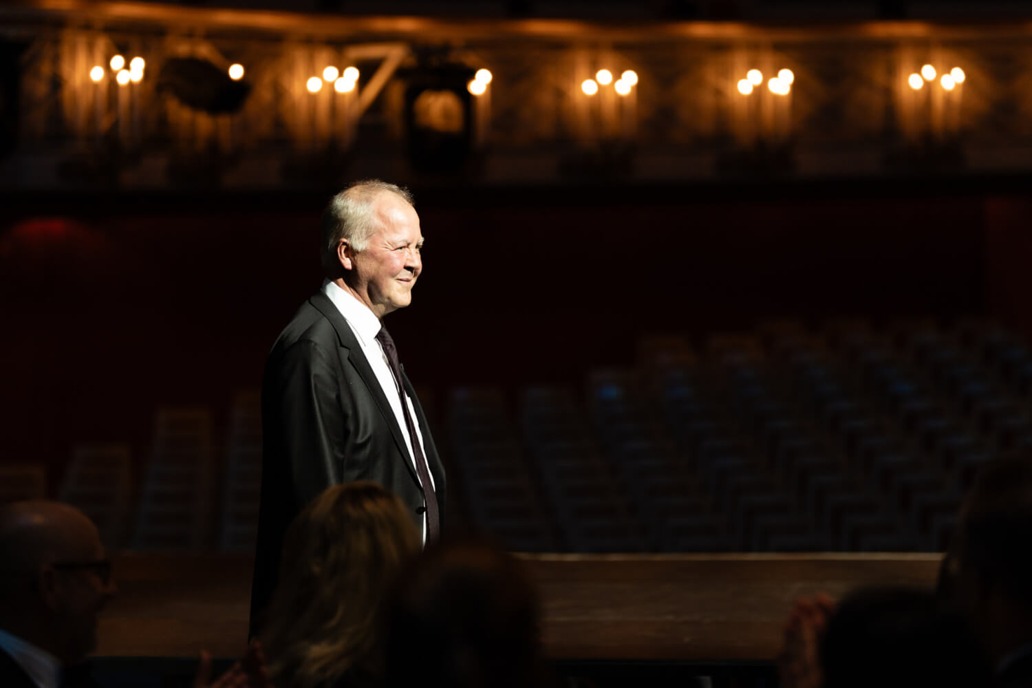 Zu sehen ist Roland Schwab in einem schwarzen Anzug mit weißem Hemd und schwarzer Krawatte. Seitlich fotografiert in einem dunklen Raum, der nur von einzelnen warmen Lichtpunkten erhellt ist.