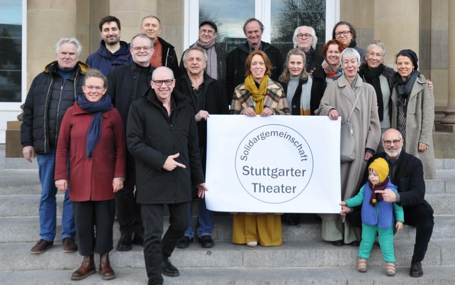 Solidargemeinschaft Stuttgarter Theater. Eine Gruppe von 18 Erwachsenen und einem Kleinkind steht auf Treppenstufen und halten ein Plakat auf dem in schwarzer Schrift steht Solidargemeinschaft Stuttgarter Theater