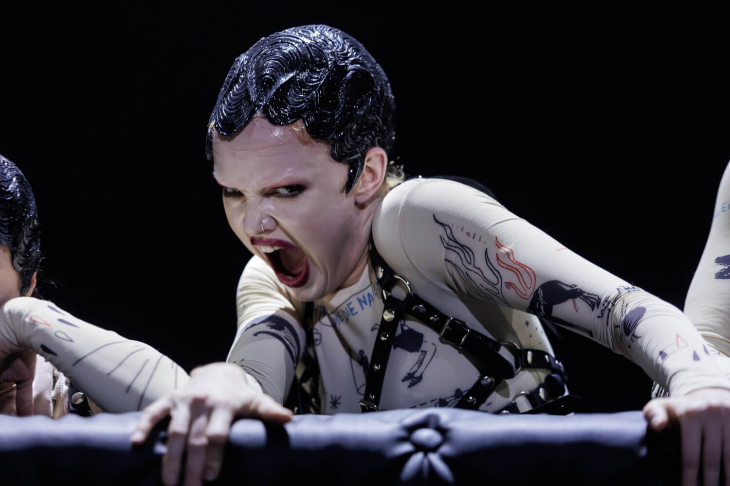 Inara Wheeler steht an einer Art Stange. Ihre beiden Hände sind fest darauf gelegt. Sie selbst hat ihren Mund zum aggressiven Schrei aufgerissen und blickt wütend nach vorn.