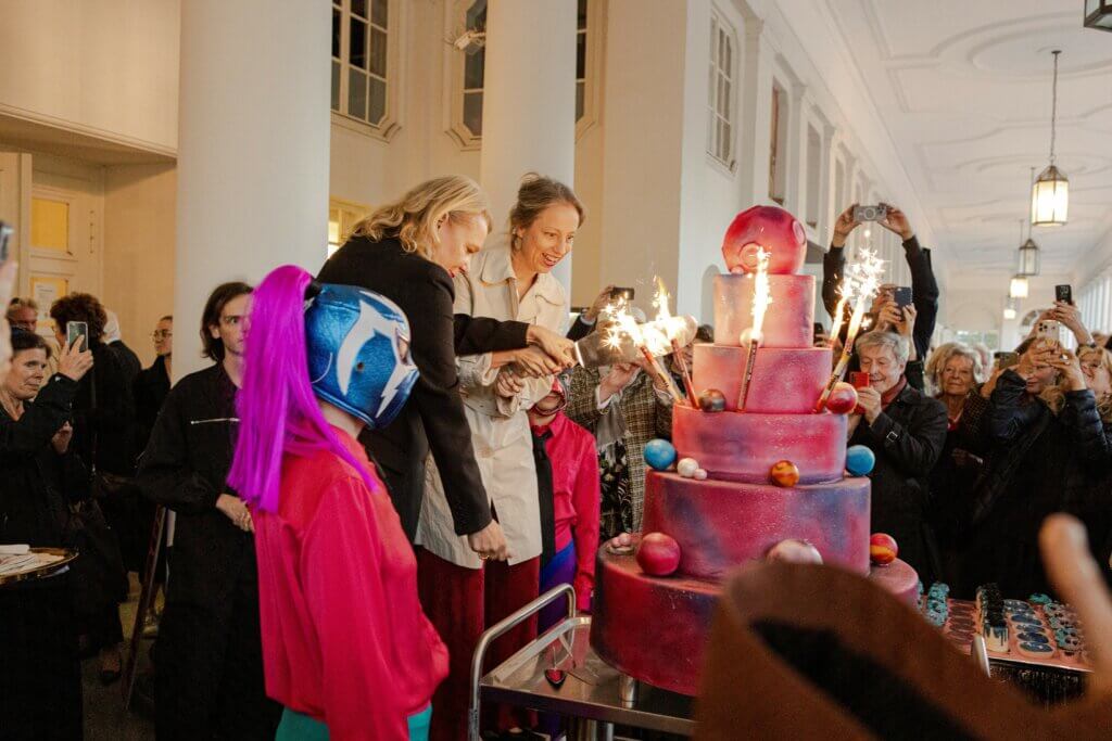 Dorothea Hartmann und Beate Heine schneiden mit einem großen Messer eine sechsstöckige bunte Torte an, an der Wunderkrzen brennen. Drumherum stehen viele Menschen, die mit ihren Smartphones filmen und fotografieren.