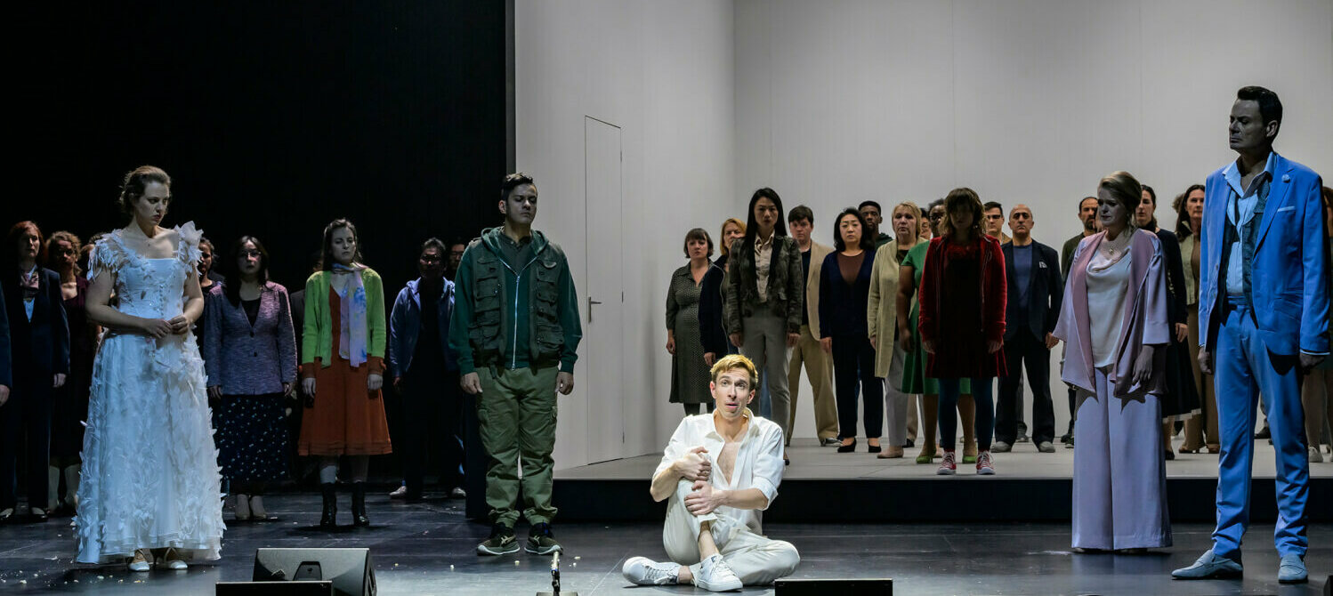 Eine darstellende Person in Weiß im Vordergrund, dahinter Ensemble und Chor