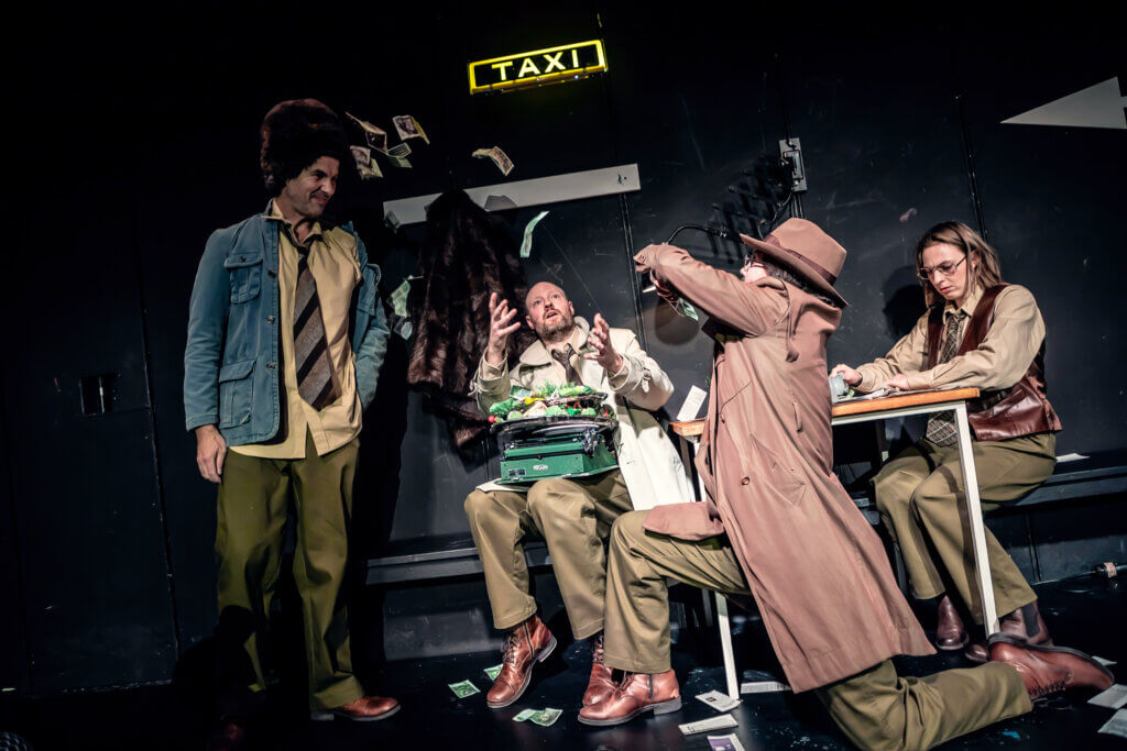 Aufführungsfoto von „Taxi nach Drüben“ von Philipp Löhle am Theater Ulm. Zwei Personen sitzen an einem Tisch sich gegenüber. Vor ihnen kniet eine andere Person, die Geldscheine in die Höhe wirft. Der Mann links am Tisch greift mit seinen Händen danach. Eine vierte Person schaut zu.