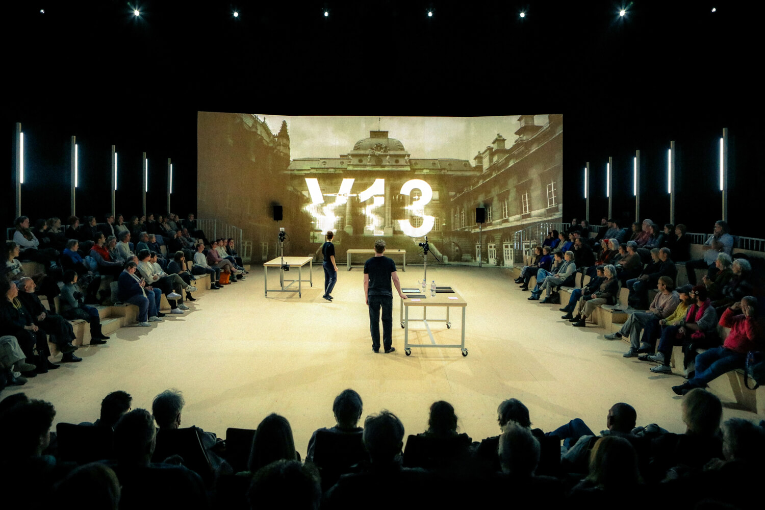 eine Bühne mit zwei Seitentribühnen mit Zuschauern. Auf die Rückwand projiziert steht V13