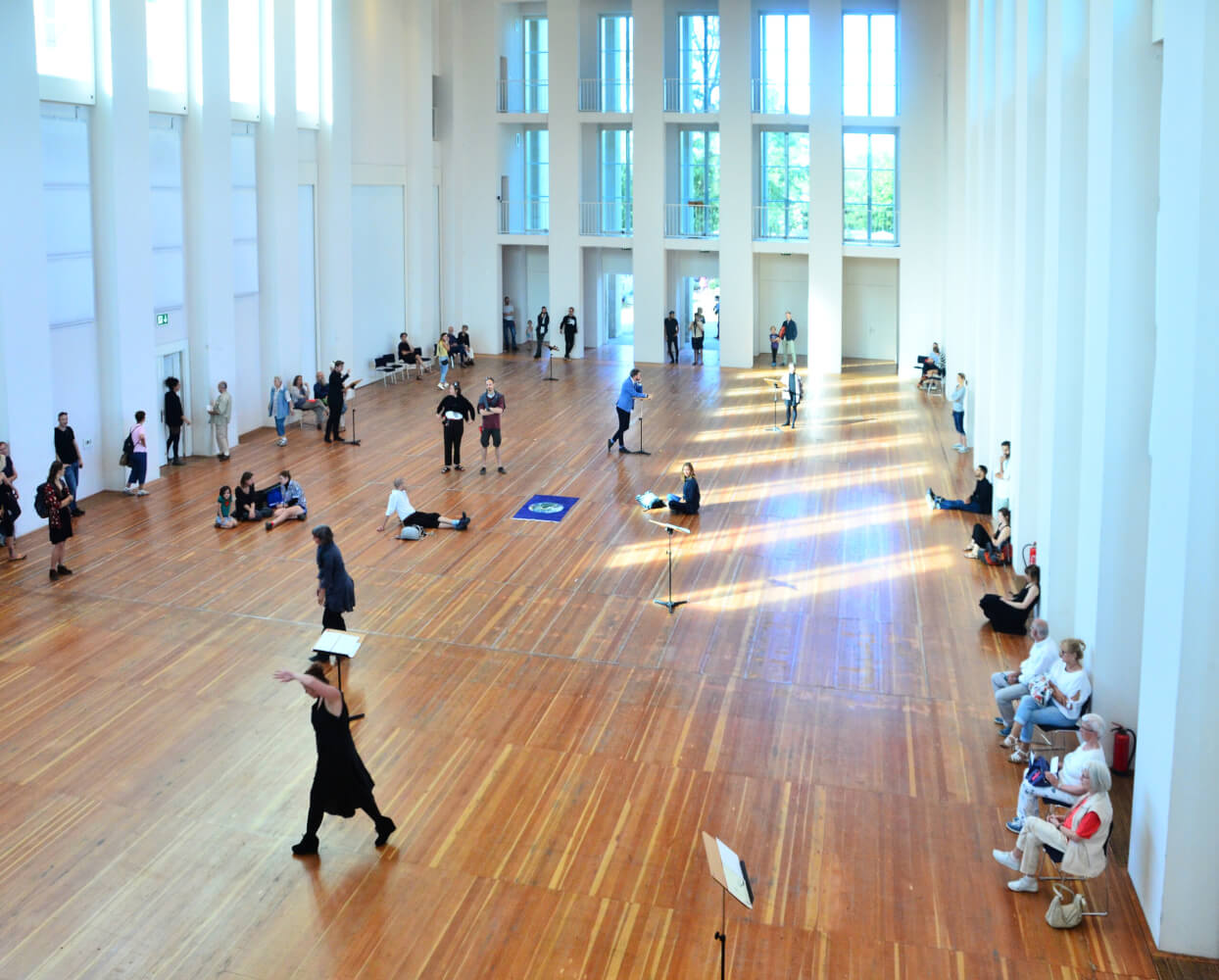 Festspielhaus HELLERAU. Ein großer offener, heller Raum mit hohen weißen Wänden, auf der Kopfseite drei Etagen mit Fenstern. Holzboden, auf dem vereinzelt Menschen stehen, sitzen und liegen.