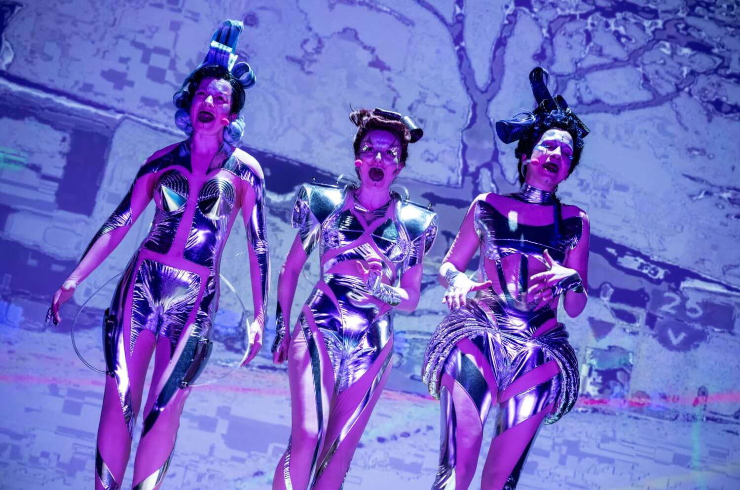 Aufführungsfoto von „KI essen seele auf (Orpheai)“ von Thomas Köck am Schauspiel Stuttgart. Drei Frauen in silbernen, durchbrochenen Ganzkörperanzügen stehen nebeinander vor einer Leinwand.