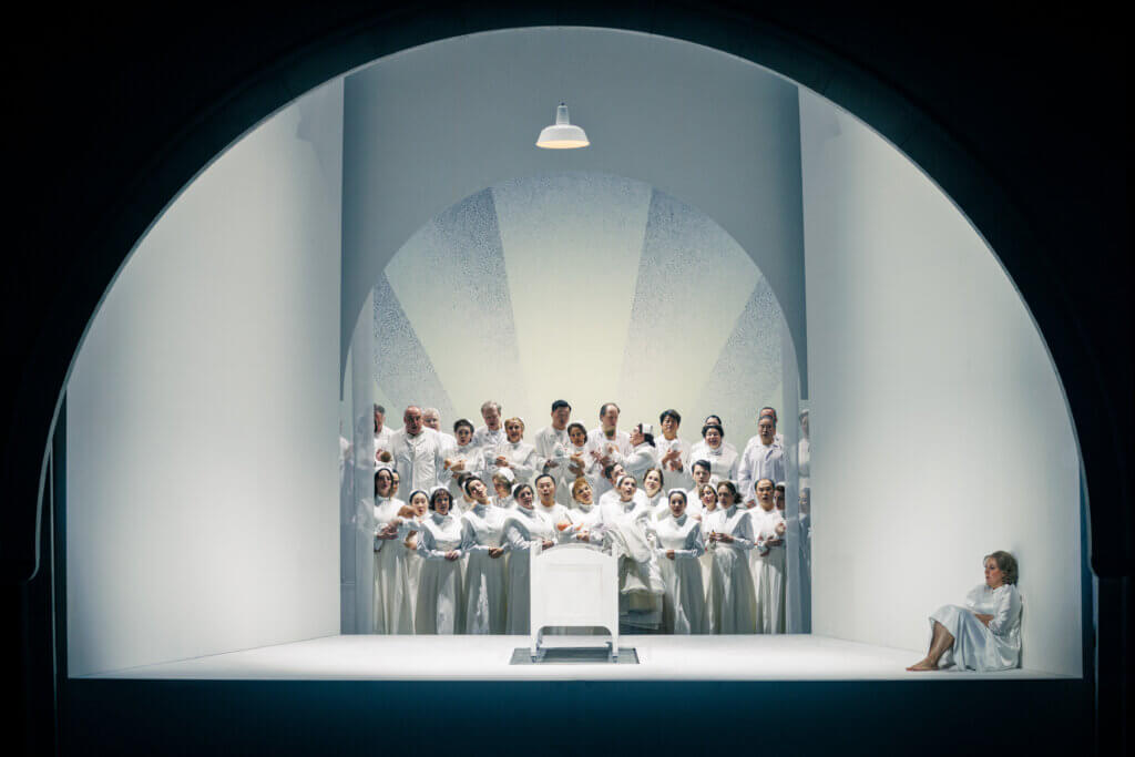 ein weißer kapellenartiger Raum mit einer Wiege und einer sitzenden Darstellerin, dahinter der Chor, alles ist weiß
