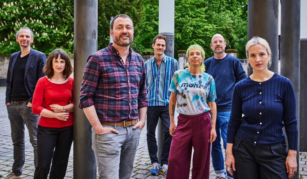 Leitungsteam des schauspiel erlangen. Sieben Menschen stehen nebeneinander, vor, neben und zwischen dunklen Säulen.