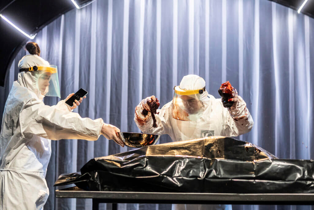 Aufführungsfoto von „√My – Episode II“ am Theater Gera. Eine Frau und ein Mann in weißen Ganzkörperschutzanzügen untersuchen den Inhalt einer schwarzen Plastiktüte auf einem Edelstahltisch. Sie hält in einer Hand ein Handy und in der anderen eine Schüssel. Er hält blutverschmierte Gegenstände in den Händen.