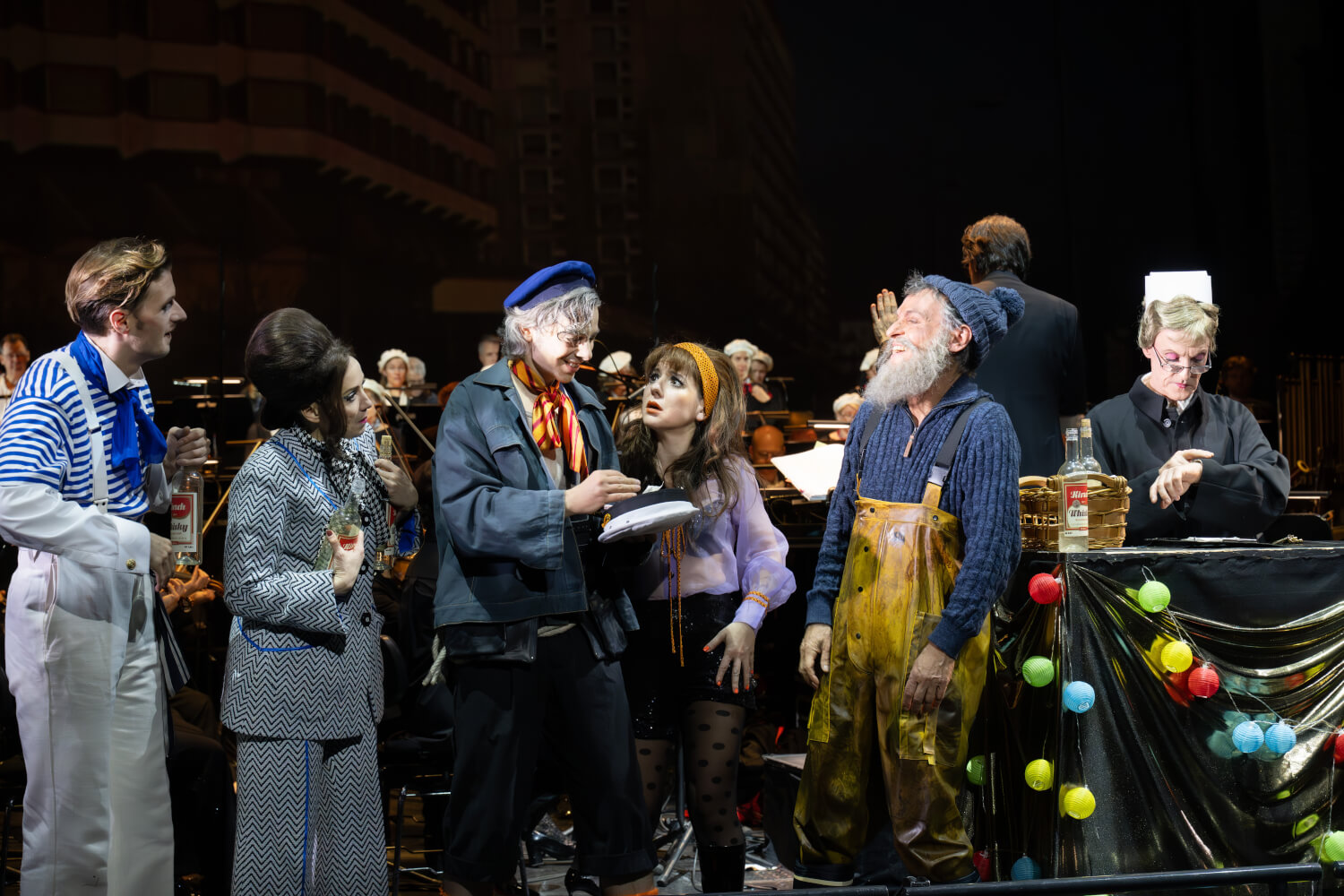 Das Ensemble steht vorne und schaut auf einen Darsteller in seiner Mitte in Seemanns-Kleidung. Im Hintergrund spielt das Orchester.
