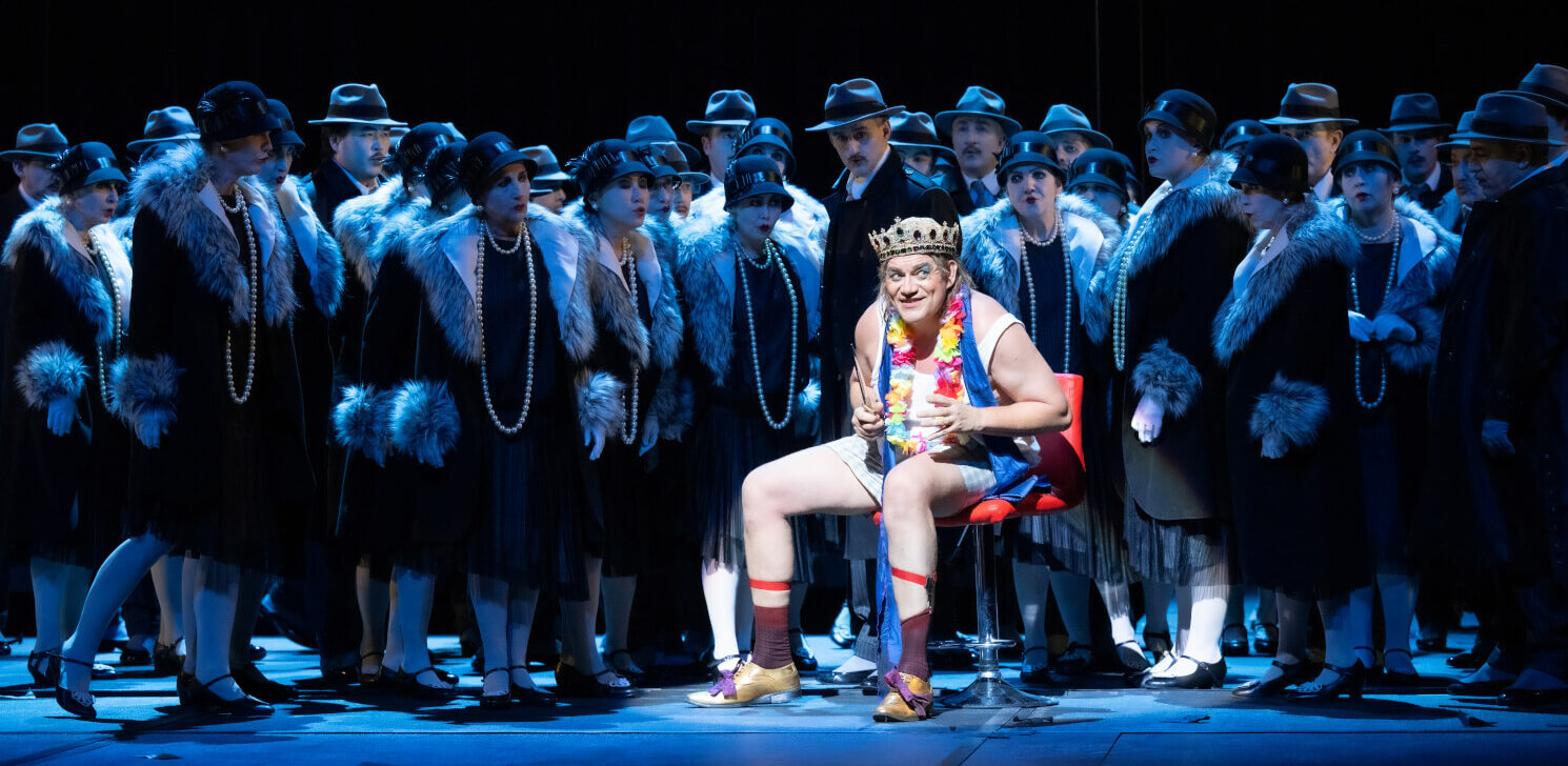 Heiko Trinsinger (Cardillac) sitzt im Scheinwerferlicht vorne in der Mitte. Im schattigen Hintergrund steht der Opernchor des Aalto-Theaters.