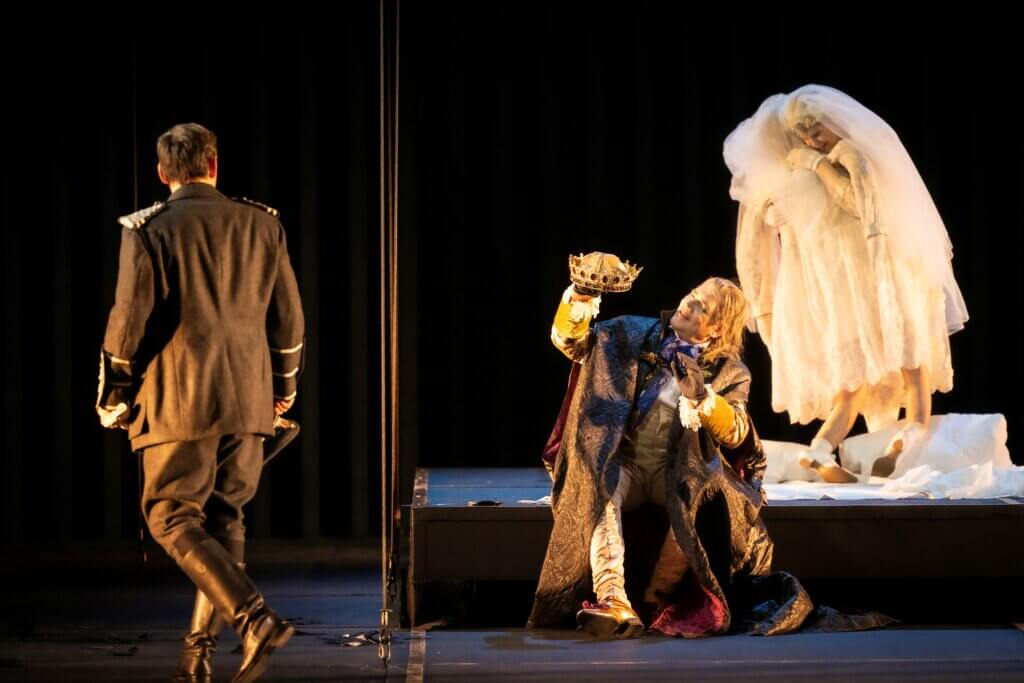 Andreas Hermann (Offizier) geht mit dem Rücken zum Publikum auf der linken Seite davon. Heiko Trinsinger (Cardillac) kniet auf dem Boden und betrachtet eine Krone. Betsy Horne (Tochter) steht im Hintergrund auf der rechten Seite.