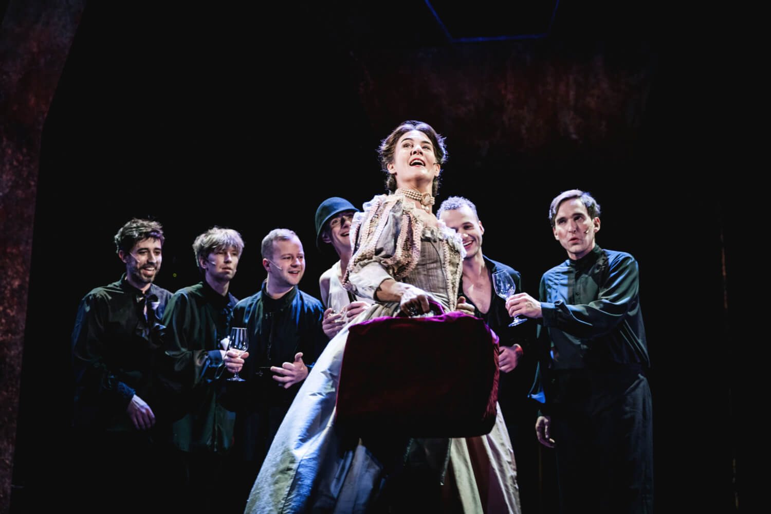 Aufführungsfoto von „Schloss der Frauen“ am Schlosstheater Celle. Eine Frau im barocken Kleid steht mit einem kleinen Koffer in der Hand vor einer Gruppe von sechs Männern, die sie anschauen.