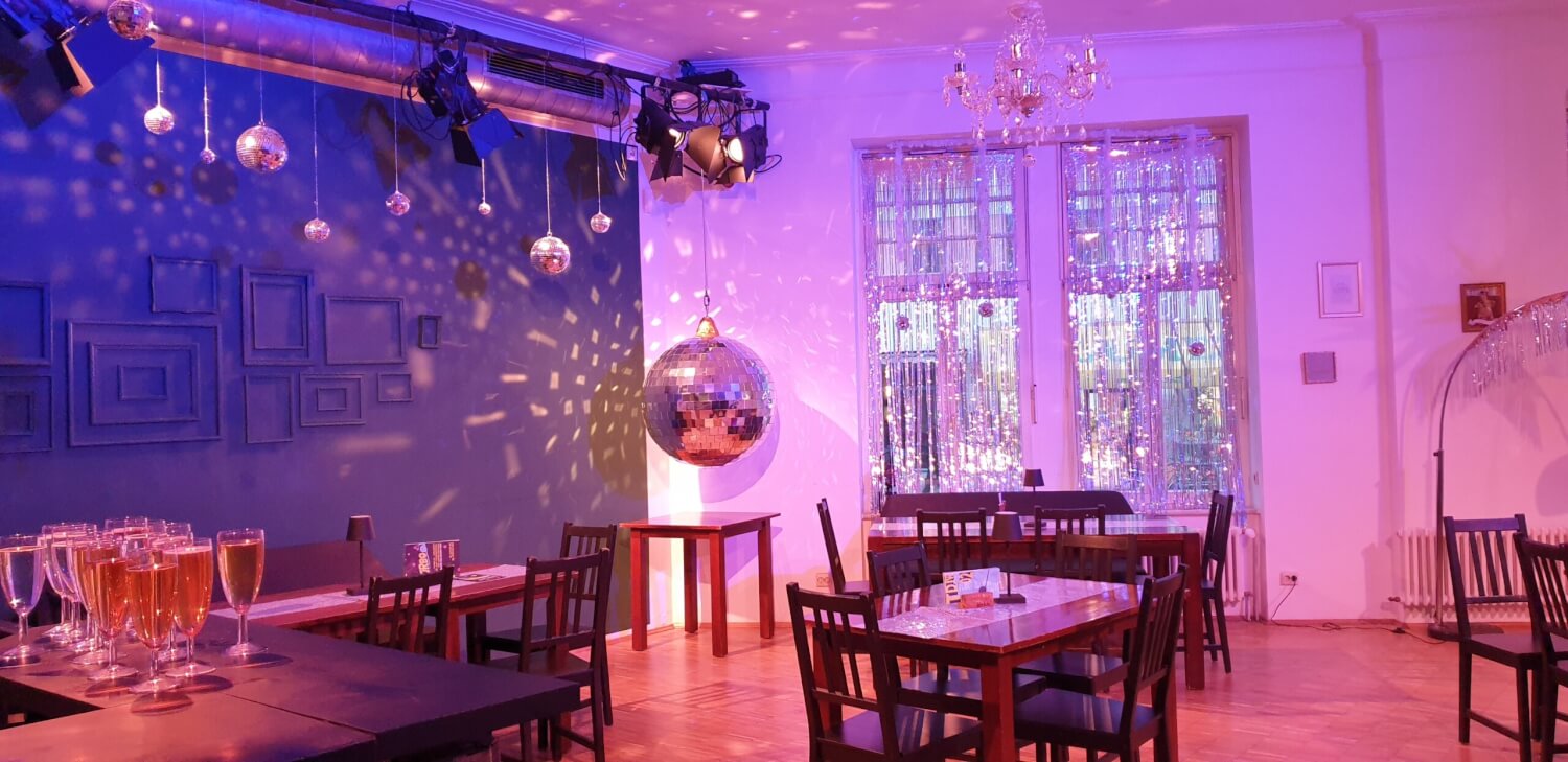 Innenraum des Theater der jungen Welt. Tische und Stühle, links vorne eine Theke mit vollen Sektgläser. In der Mitte eine mit glitzernden Fäden geschmücke Fensterfront. In der linken Ecke eine große glitzernde Discokugel.