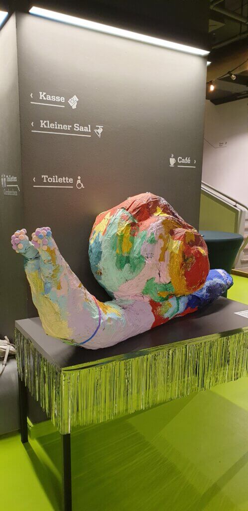 Auf einem Tisch steht eine Pappmaché-Schnecke, die mit bunten Flecksen bemalt ist. Der Boden ist grün und hinter der Schnecke sind Wegweiser zu sehen.