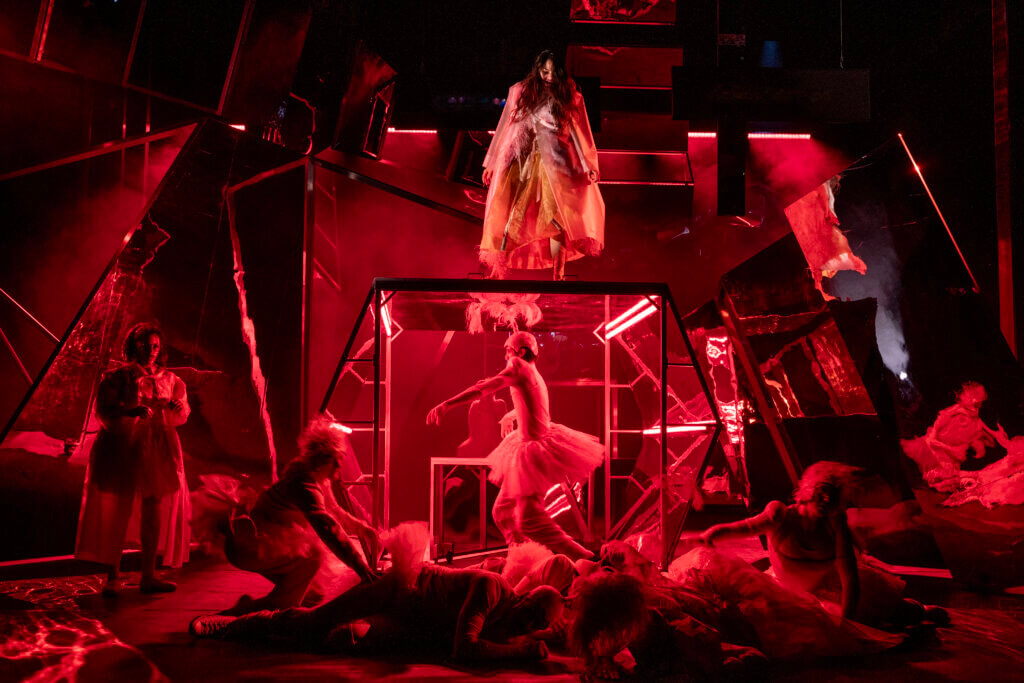 Auf der Bühne herrscht eine bedrohlische Stimmung. Das Ensemble liegt im Raum verteilt. Eine Person scheint wie in einem Hamsterrad zu laufen.