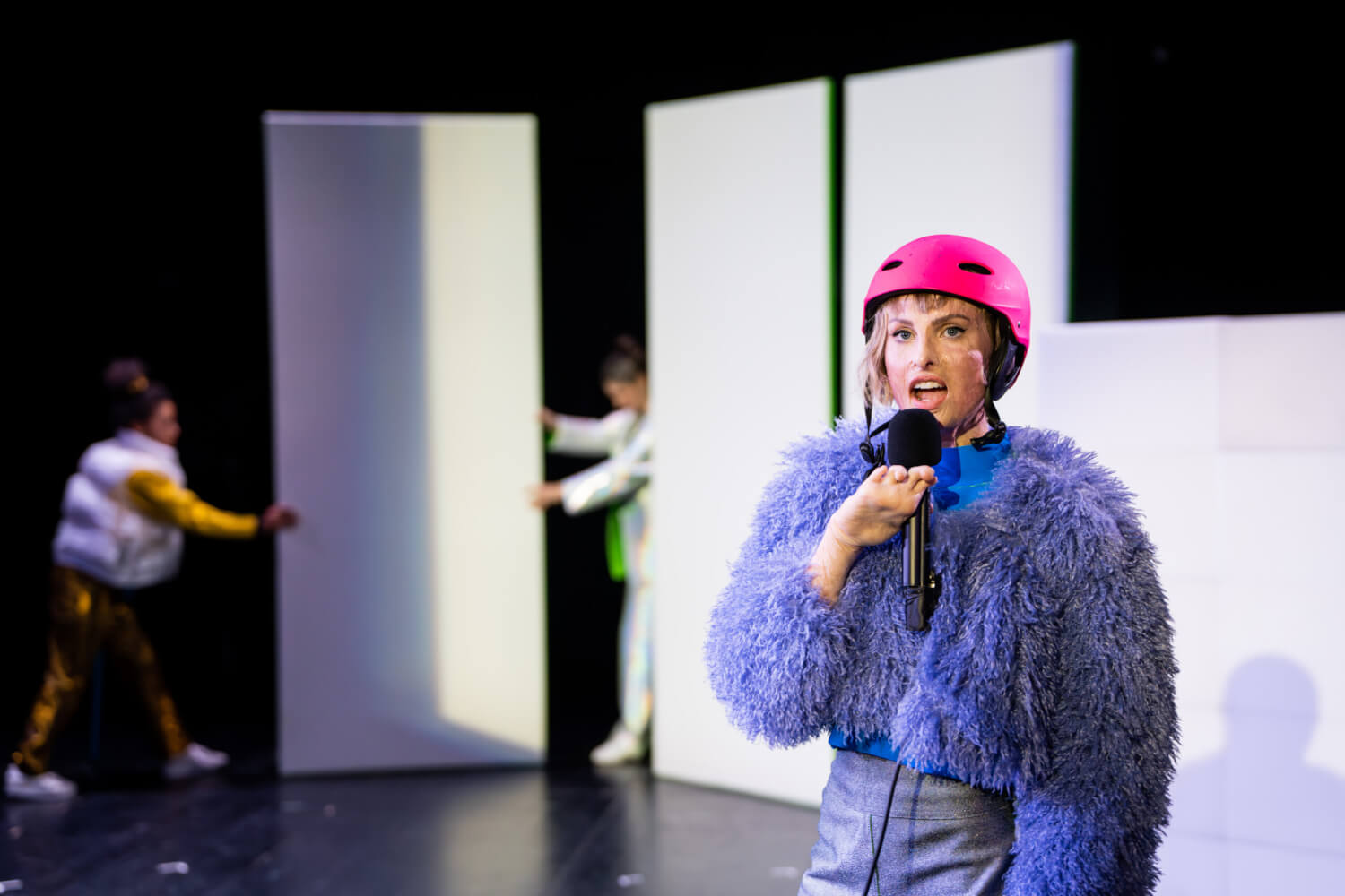 Szenenfoto aus „ZERBRECH-LICH“. Eine Performerin mit einem Mikrofon in der Hand und einem pinken Fahrradhelm steht im Vordergrund. Im Hintergrund schieben zwei Frauen eine Stellwand an zwei andere Stellwände