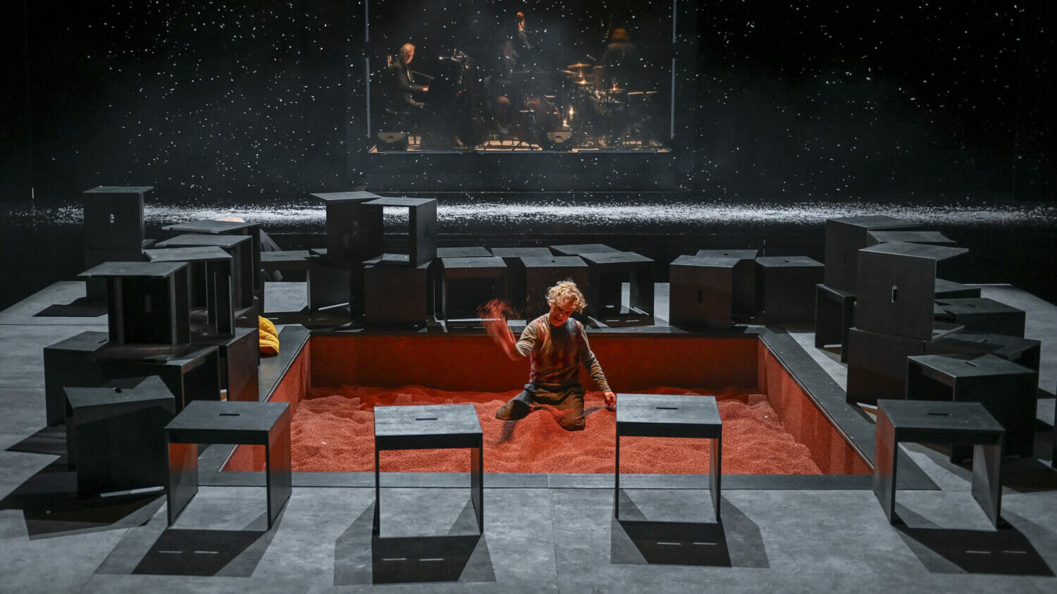 In der Mitte der Bühne ist in den Boden ein Sandkasten eingelassen. Der Sand ist nicht sandfarben sondern rot. Dort sitzt Alfi und spielt.