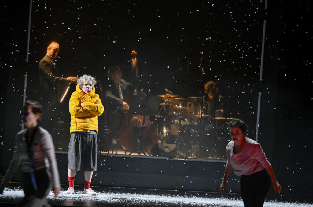 Zwei Darstellende bewegen sich von rechts nach links. Einer steht dick in eine Jacke gekleidet und starrt nach vorn. Im Hintergrund ist die Band zu sehen. Es schneit.