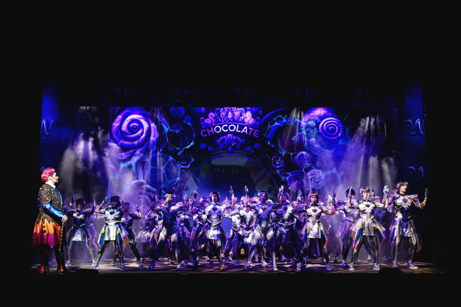 Aufführungsfoto von „Charlie und die Schokoladenfabrik“ von Roald Dahl am Theater Regensburg. Ein Mann im bunten Kostüm steht links an der Seite. Lilafarbenes Licht strahlt eine verzierte Wand an, auf der Chocolate steht. Davor steht eine große Gruppe von Menschen, die die Arme hoch strecken.