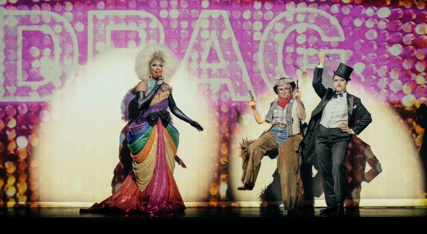 Links steht eine Dragqueen mit Mikrofon. Rechts posen zwei Darstellende mit erhobenem linkem Bein.