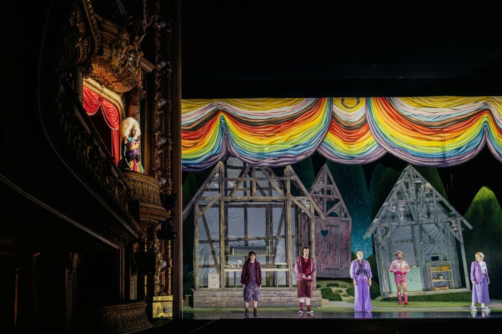 Vier Ensemblemitglieder stehen verteilt vor Fachwerkhäusern. Über ihnen hängen runde Vorhänge in Regenbogenfarben-