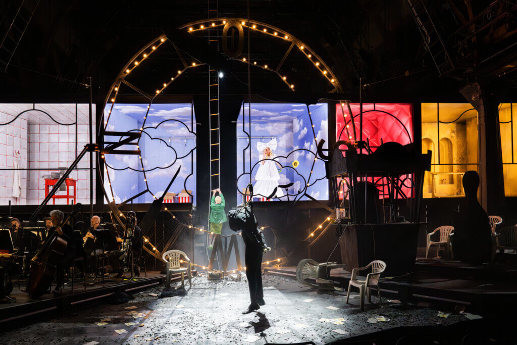 eine Bühne, im Vordergrund hängt ein Darsteller, dahinter erleuchtete Guckkastenraumbühnen