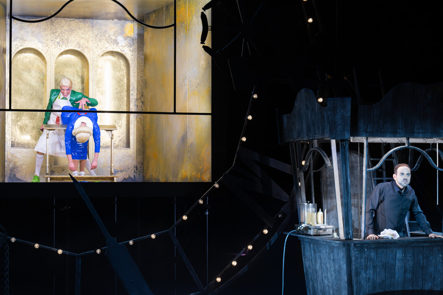 hinten zwei Darstellende in grellen Kostümen, vorne in einem kleinen Häuschen einer in Schwarz