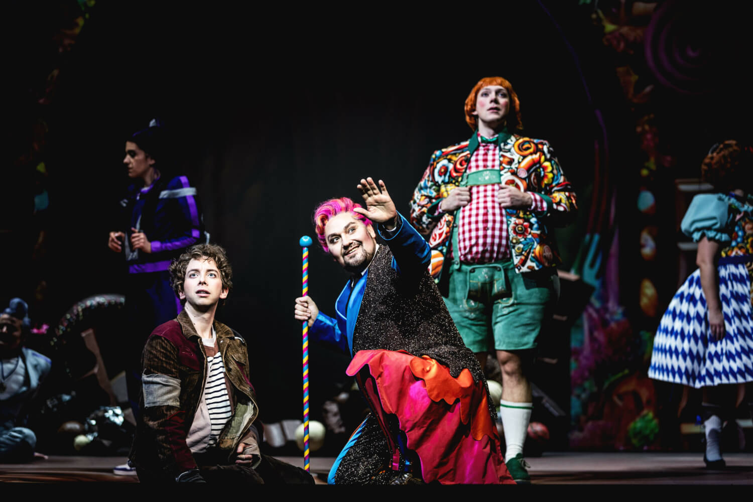 Aufführungsfoto von „Charlie und die Schokoladenfabrik“ von Roald Dahl am Theater Regensburg. Ein Mann in buntem Kostüm und einer Zuckerstange als Gehstock, kniet neben einem Jungen. Beide schauen gespannt nach vorne. Im Hintergrund ein Mann in grüner Lederhose und rot kariertem Hemd schaut auch in dieselbe Richtung.