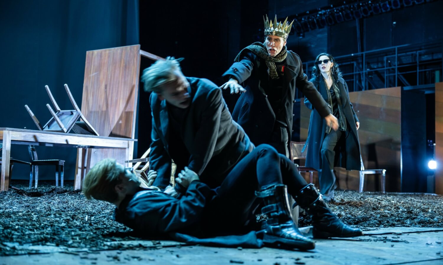 Aufführungsfoto von „Hamlet“ von William Shakespeare am Schauspiel Stuttgart. Hamlet stürzt sich auf den am Boden liegenden Laertes. Claudius mit einer Krone auf dem Kopf stürzt zu den beiden. Im Hintergrund Gertrud mit schwarzem Mantel und dunkler Sonnenbrille.