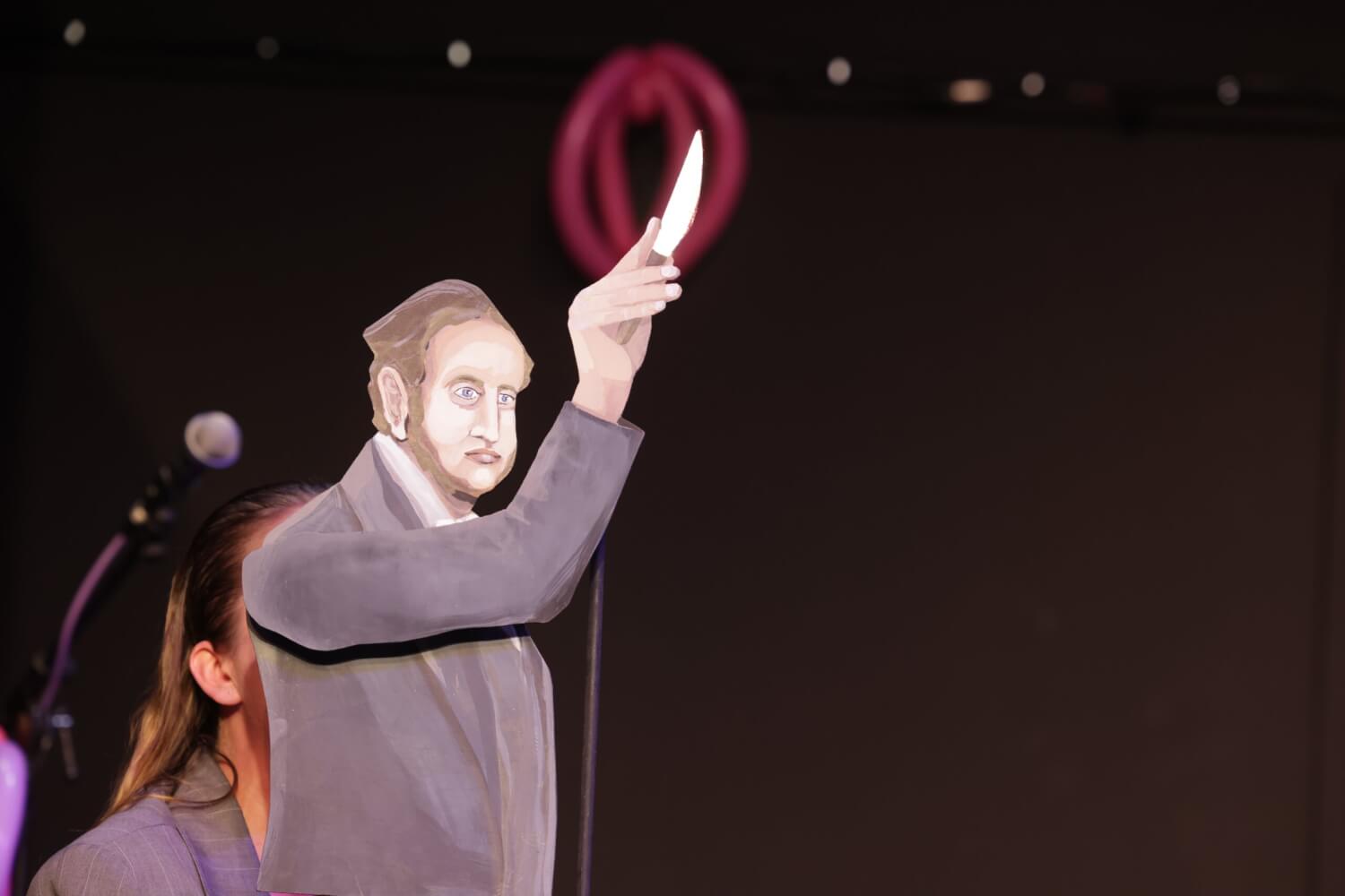 Aufführungsfoto von „Der Ursprung der Welt“ an der Neuen Bühne Senftenberg. Eine Frau hält eine Stabpuppe, die ein Messer in der Hand hält.