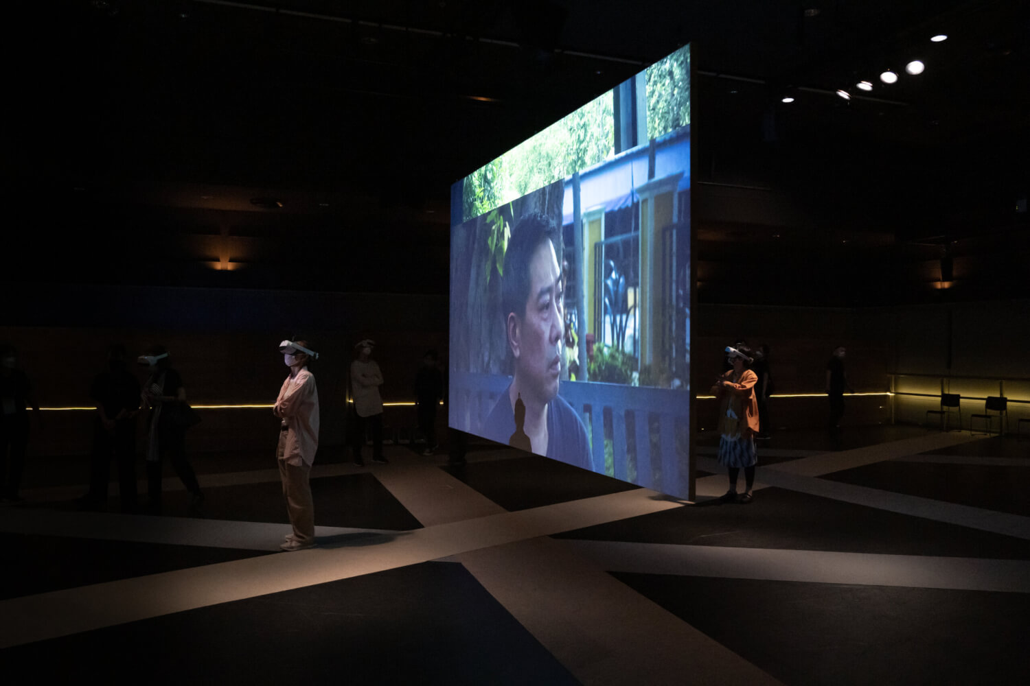 Aufführungsfoto von „A Conversation with the Sun (VR)“ von Apichatpong Weerasethakul. In der Mitte eine große Leinwand, auf der ein Mann bis zu den Schultern zu sehen ist. Davor uns dahinter stehen zwei Menschen, die VR-Brillen tragen und mit verschränkten Armen auf der Bühne stehen.