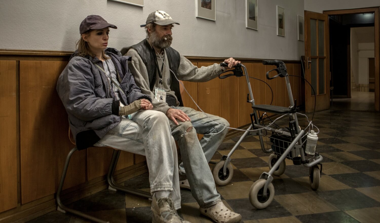 Signa und Arthur Köstler in „Das halbe Leid“ 2017 am Deutschen Schauspielhaus Hamburg. Eine Frau und ein Mann sitzen nebeneinander in einem Flur. Über ihnen Bilder. Neben dem Mann ein Rollator.