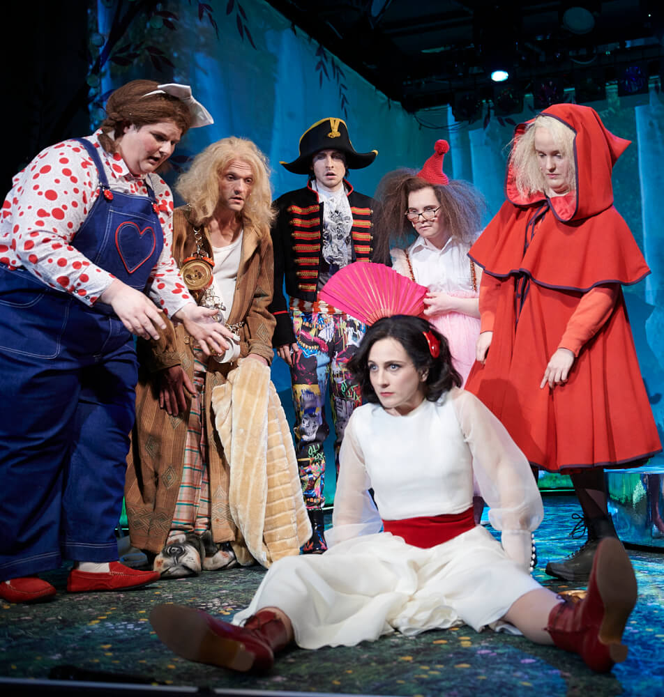 Aufführungsfoto von „Schneewittchen“ von Studiyou, inklusives Studio für Schauspieler:innen am Schauspiel Wuppertal. Schneewittchen sitzt breitbeinig auf dem Boden. Hinter ihr stehen fünf Figuren in verschiedenen Kostümen und schauen sie an.