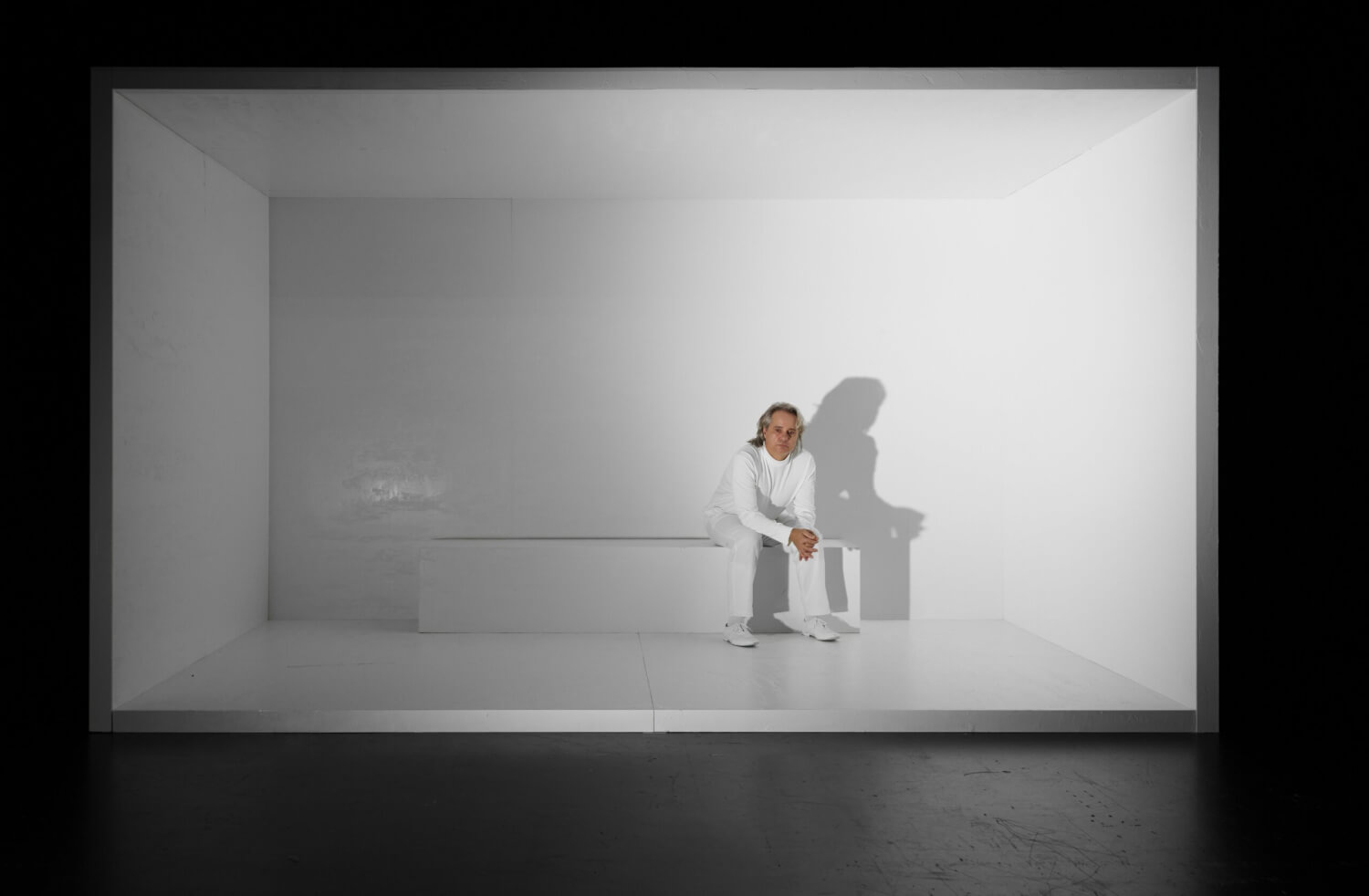 Aufführungsfoto von „Serotonin“ nach Michel Houellebecq am Hans Otto Theater Potsdam. Ein Mann ganz in weiß sitzt auf einem langgezogenen, weißen Quader in einem sonst leeren weißen Raum.