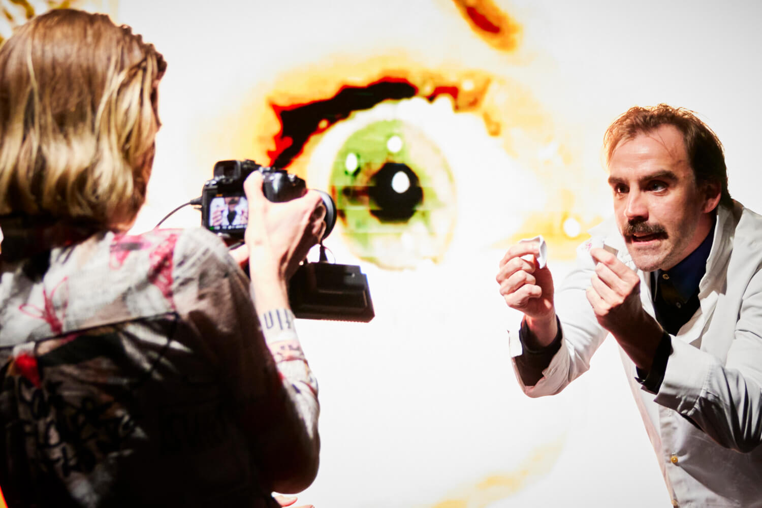 Aufführungsfoto von „Mythos P.A.N.“ im XRT am Staatstheater Nürnberg. Eine Frau links hält eine Kamera und filmt auf einen Mann, der zwei kleine Gegenstände in die Kamera hält.